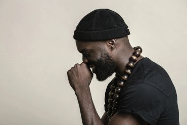 Profile of a pensive man wearing a black beanie and wooden bead necklace.